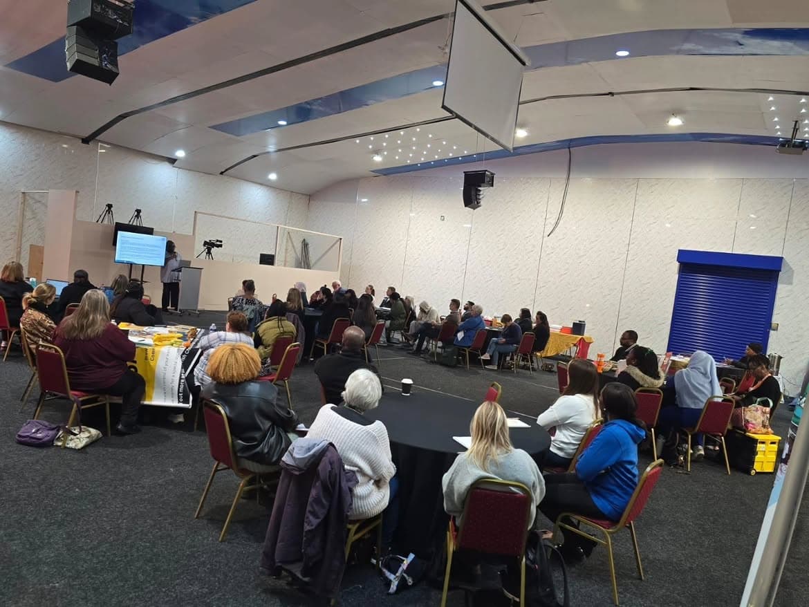 A community hall with around forty people seated at round tables, facing a speaker presenting from a screen at the front.
